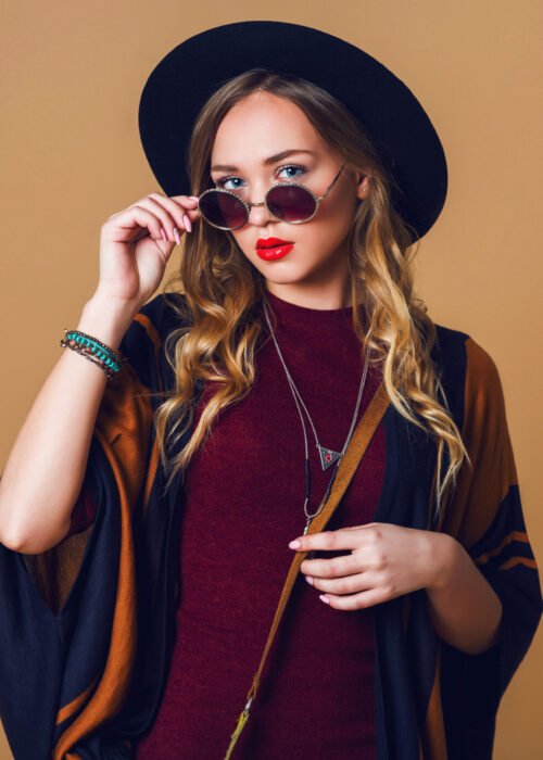 Studio close up portrait of young fresh blonde  woman in brown straw poncho , wool black trendy hat and round glasses looking at camera. Green leather had bag.