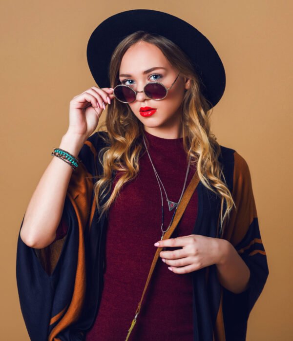 Studio close up portrait of young fresh blonde  woman in brown straw poncho , wool black trendy hat and round glasses looking at camera. Green leather had bag.